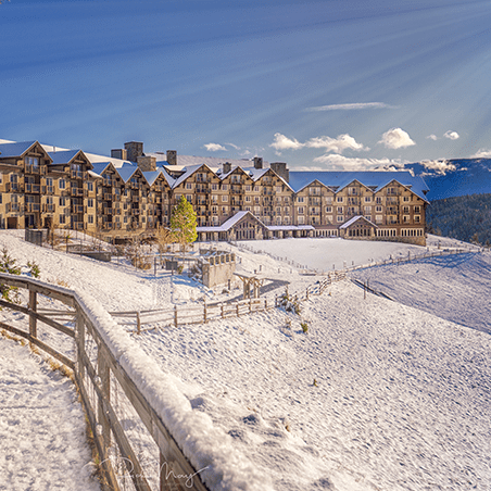 Large mountain lodge surrounded by snow-covered ground and hills, with a wooden fence and distant mountains under a clear blue sky.