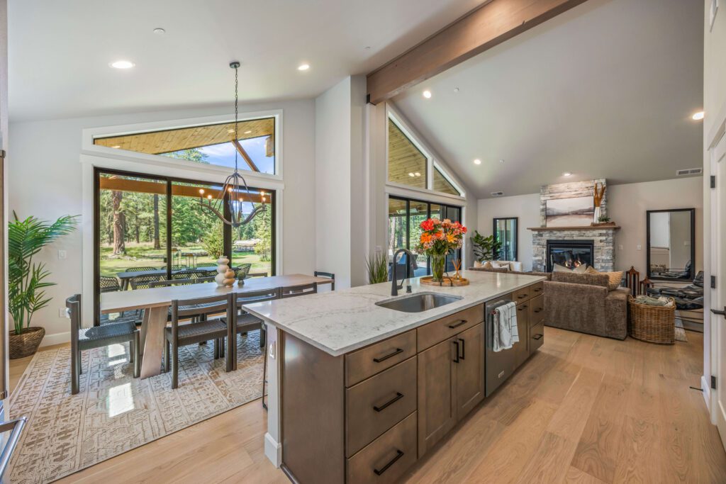Modern open-concept kitchen and dining area with marble island, wood floors, large windows, and views of a green outdoor landscape.