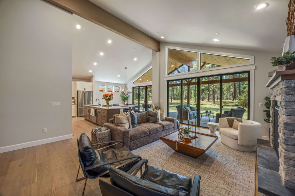 Modern open-concept living room with large windows, neutral decor, stone fireplace, and view of a wooded backyard. Kitchen and dining area are visible in the background.