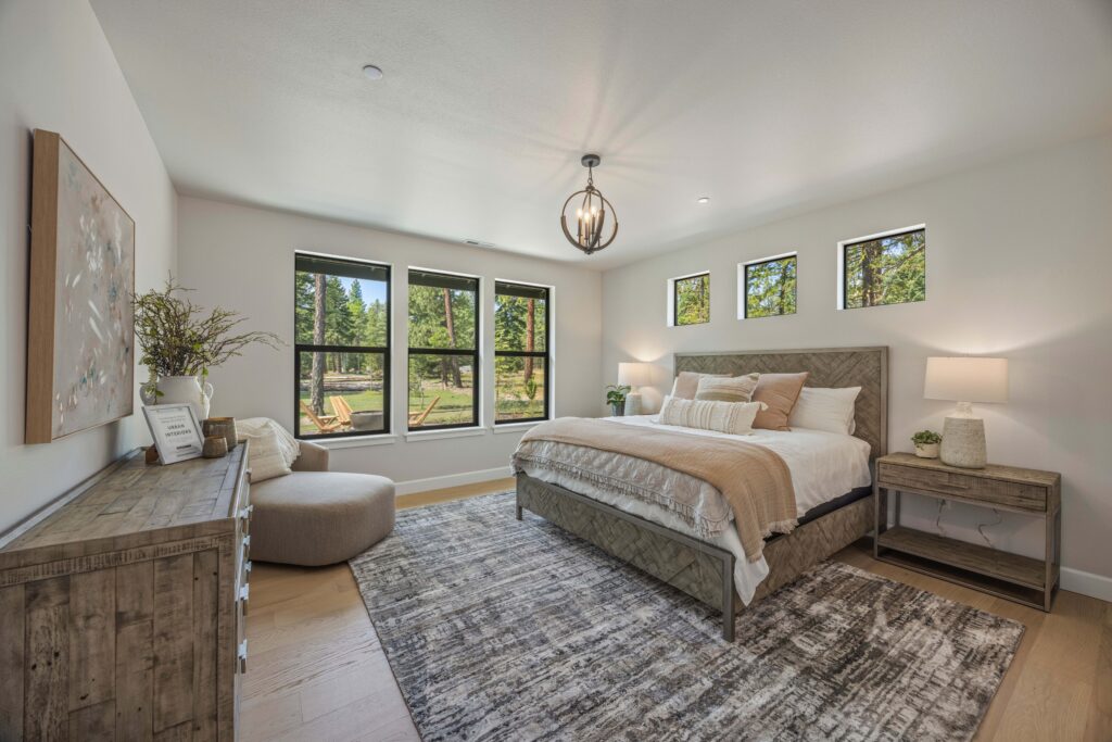 Modern bedroom with large windows, a bed with neutral bedding, two wooden nightstands with lamps, a dresser, ottoman, and a patterned rug on wooden floors.