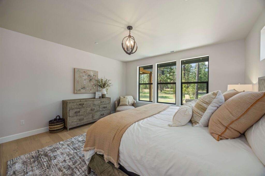 Modern bedroom with a large bed, neutral bedding, dresser, armchair, and three large windows overlooking a green outdoor area.