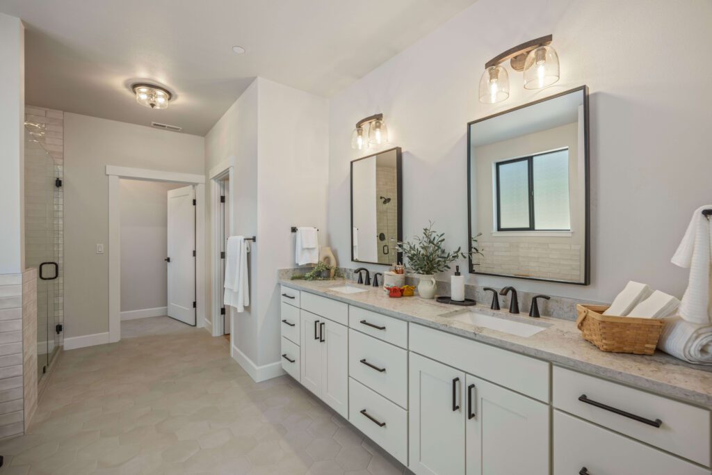Modern bathroom with double sink vanity, black fixtures, large mirrors, and a glass-enclosed shower. Towels, a small plant, and a window complete the clean, neutral decor.