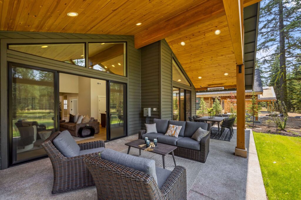 Covered patio with wicker furniture, gray cushions, coffee tables, and large glass doors, overlooking a lawn and surrounded by trees.