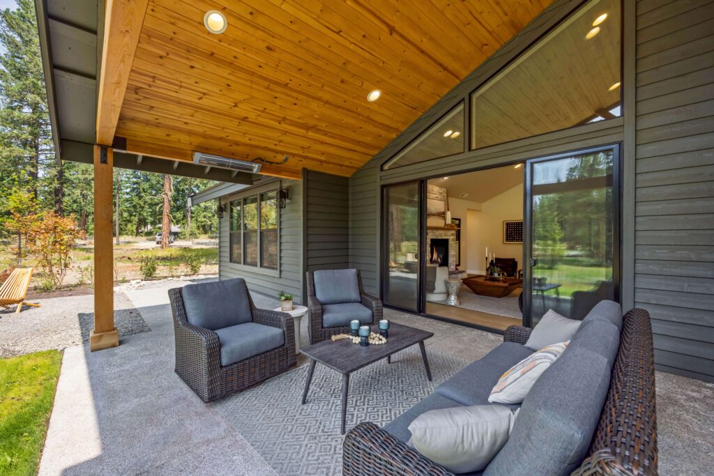 Covered patio with wicker furniture and cushions, a coffee table with candles, and sliding glass doors leading to a modern living room; surrounded by trees and greenery.