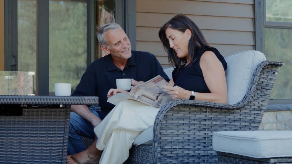 A man and woman sit together on a wicker patio sofa, drinking from mugs while the woman looks at a magazine. They are outdoors near a house.