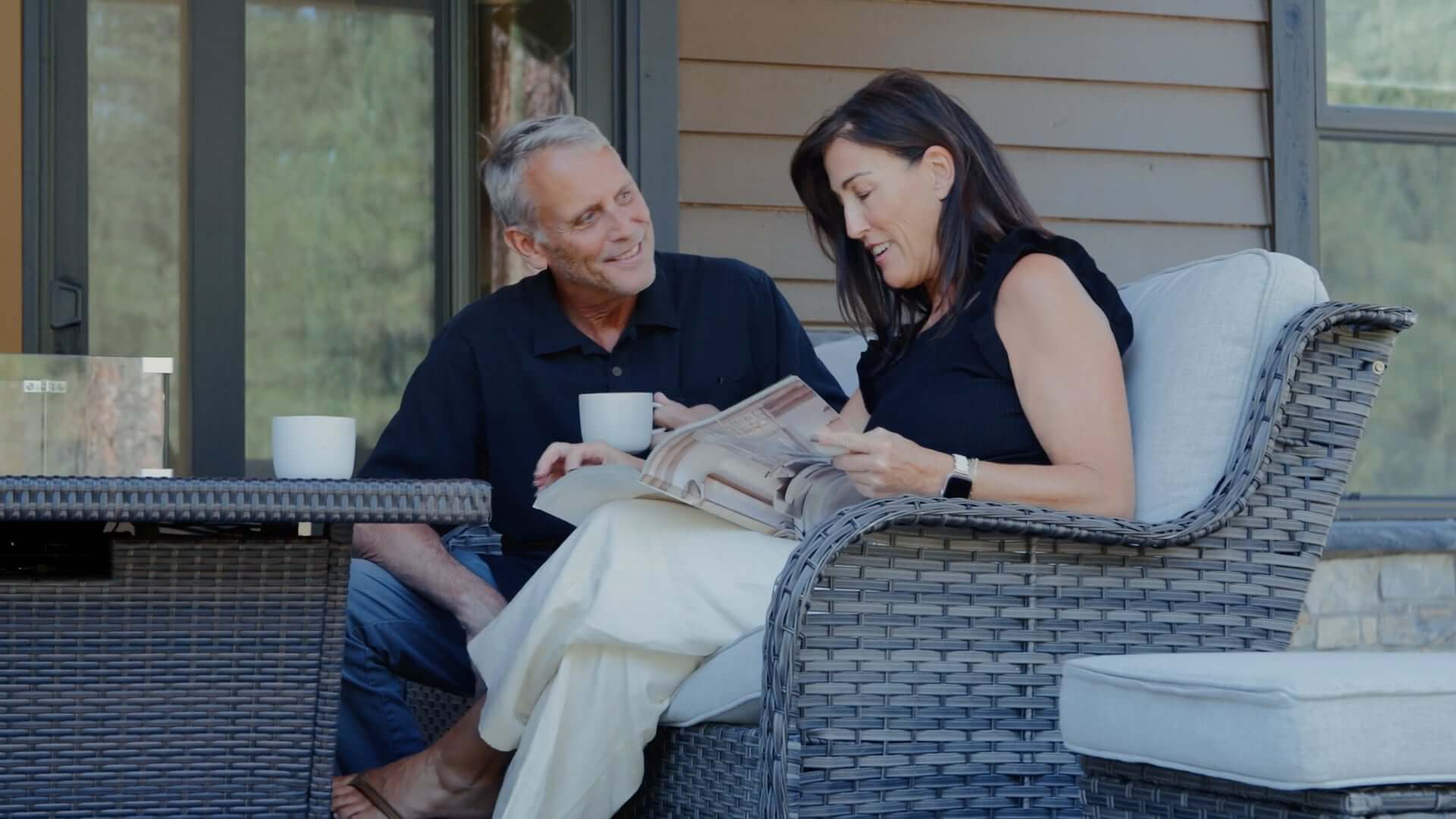 A man and woman sit together on a wicker patio sofa, drinking from mugs while the woman looks at a magazine. They are outdoors near a house.