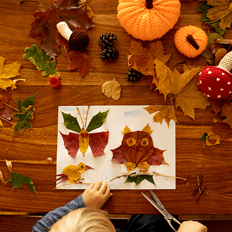A child making art with autumn leaves and paper on a wooden table, surrounded by leaves, pinecones, yarn pumpkins, and a stuffed mushroom.