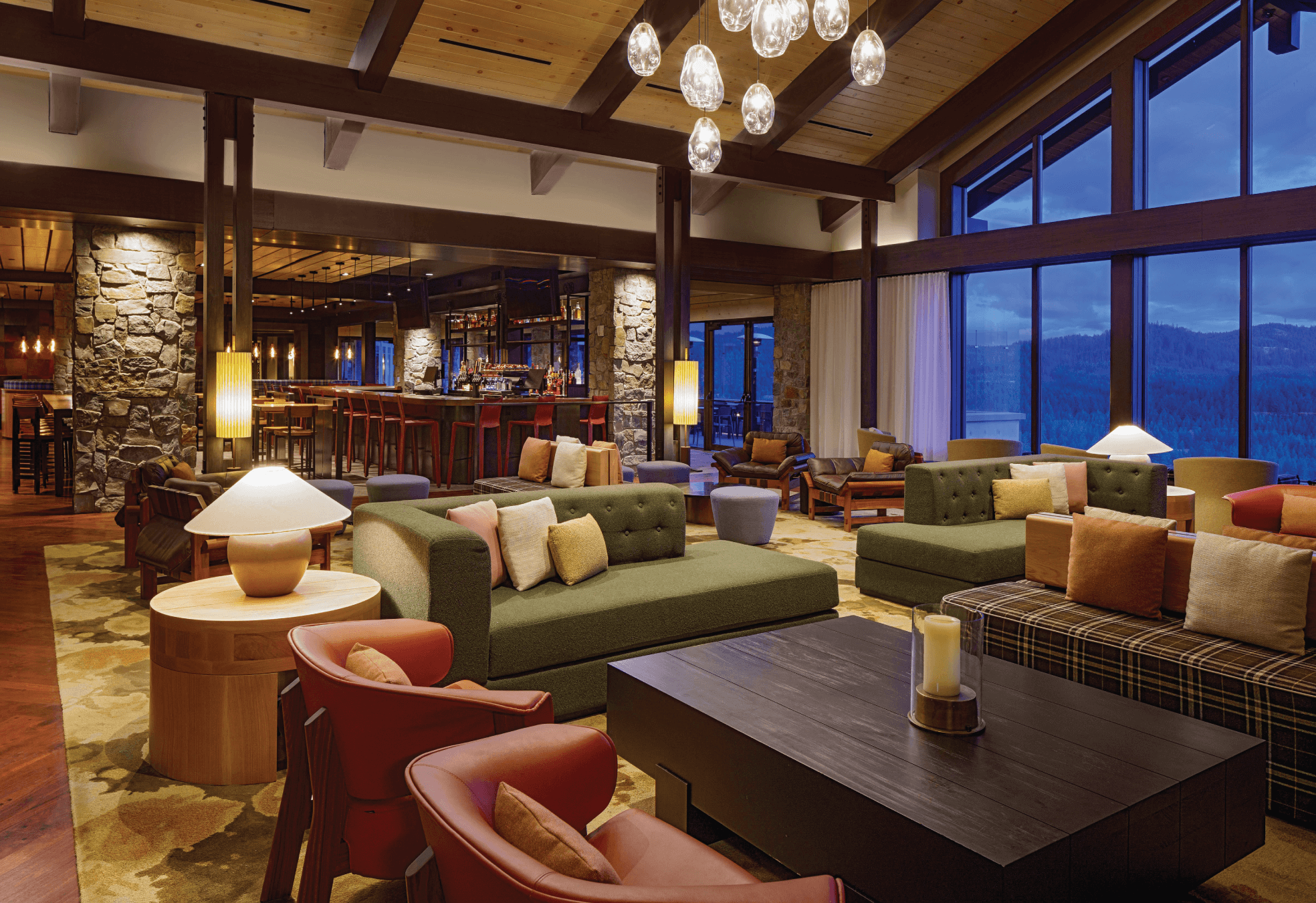 Spacious hotel lounge with modern sofas, armchairs, large windows overlooking mountains, warm lighting, and a bar area in the background.