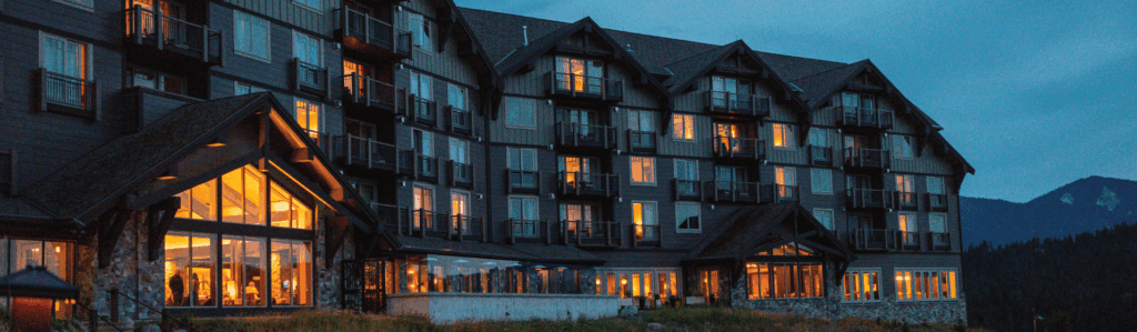A large multi-story lodge with illuminated windows at dusk, set against a mountainous backdrop.
