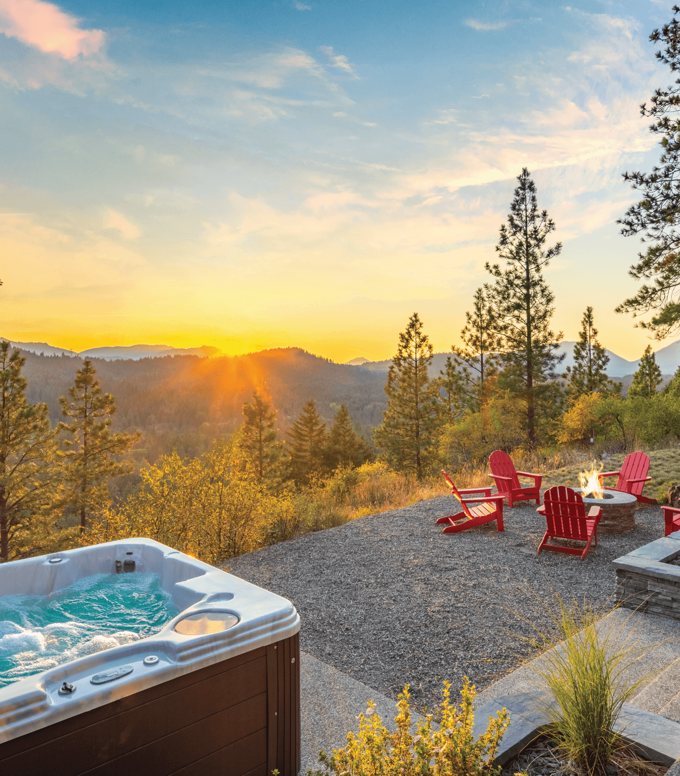 A hot tub and red Adirondack chairs surround a fire pit on a gravel patio overlooking a forested mountain landscape at sunset.