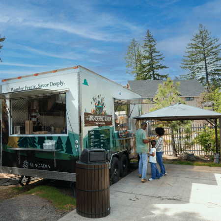 A family of three stands near a food truck called The Wandering Whisk, parked outdoors by trees and a gazebo on a sunny day. The parents are ordering while their child stands beside them.
