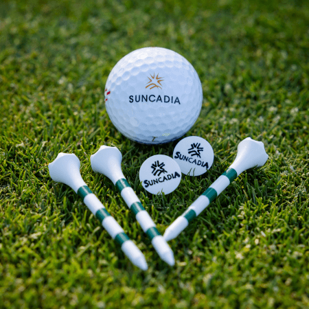 A Suncadia-branded golf ball, three golf tees, and two ball markers with the Suncadia logo are arranged on green grass.
