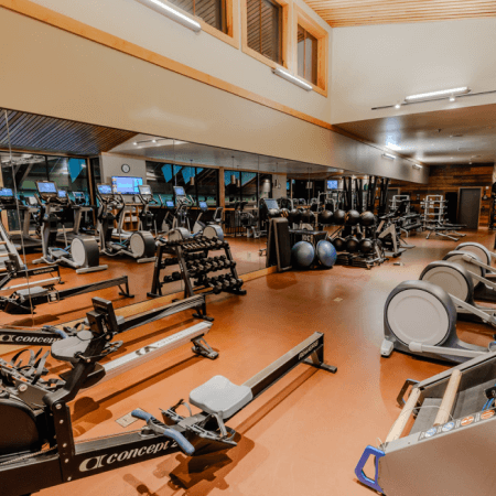 A spacious gym with rowing machines, treadmills, dumbbells, exercise balls, and other fitness equipment. Large mirrors line one wall and there are high windows letting in natural light.