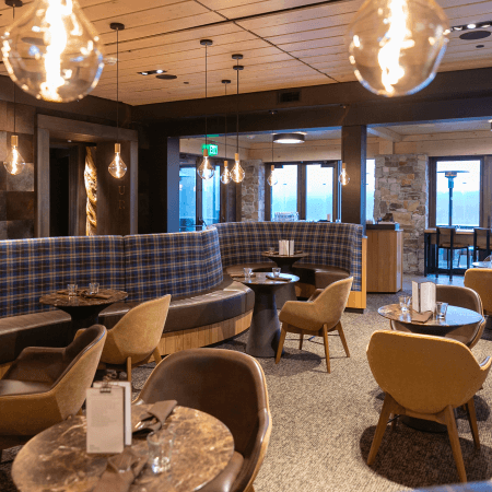 Modern restaurant interior with warm lighting, round tables, cushioned chairs, and a curved plaid booth. Glass pendant lights hang from the ceiling, and large windows reveal an outdoor view.