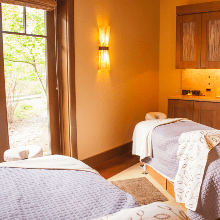 A cozy spa room with two massage tables covered in light gray quilts, soft beige towels, and pillows. Warm lighting and a large window with a leafy outdoor view create a relaxing atmosphere.