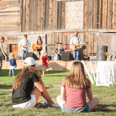 Two girls sit on grass watching a live band perform on an outdoor stage in front of a rustic wooden building, with children playing and people standing nearby. White chairs and tables are set up to the right.