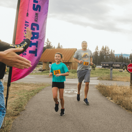 A young girl and an adult man, both wearing race bibs, run on a paved path. The girl smiles as she nears a high-five by a colorful flag. Trees and buildings are visible in the background.