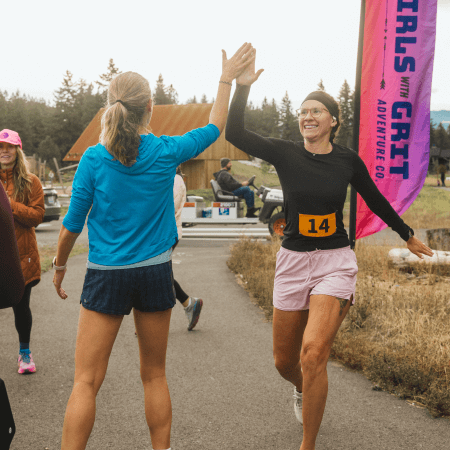 Two women high-five at the finish line of a race. One wears a blue jacket and shorts, the other wears a black top, pink shorts, and race bib 14. A Girls with Grit banner is visible in the background.