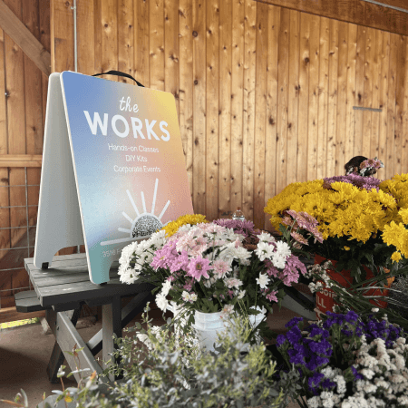 A display of colorful flowers in buckets sits on a table next to a sign that reads, The Works: Hands-on Classes, DIY Kits, Corporate Events, inside a wooden structure.