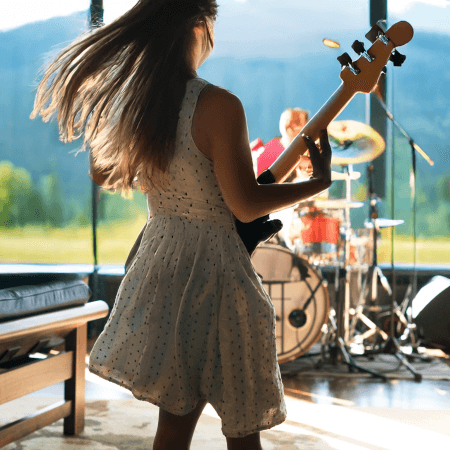 A woman in a white dress plays bass guitar with her hair swinging as she performs onstage, with a drummer in the background and large windows revealing a scenic outdoor view.