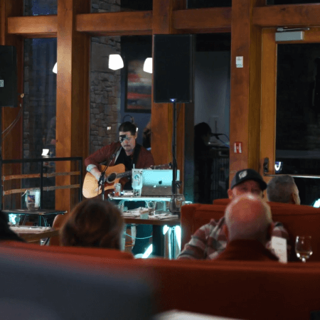 A musician in sunglasses plays an acoustic guitar and sings into a microphone on a small stage in a cozy restaurant, while several people sit at tables and listen.