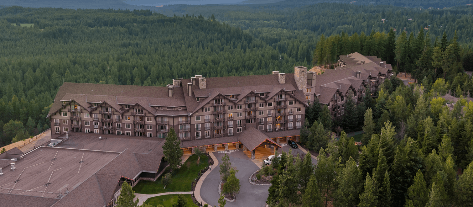 A large, multi-story lodge-style hotel surrounded by dense green forest, with mountains visible in the distance under a cloudy sky.