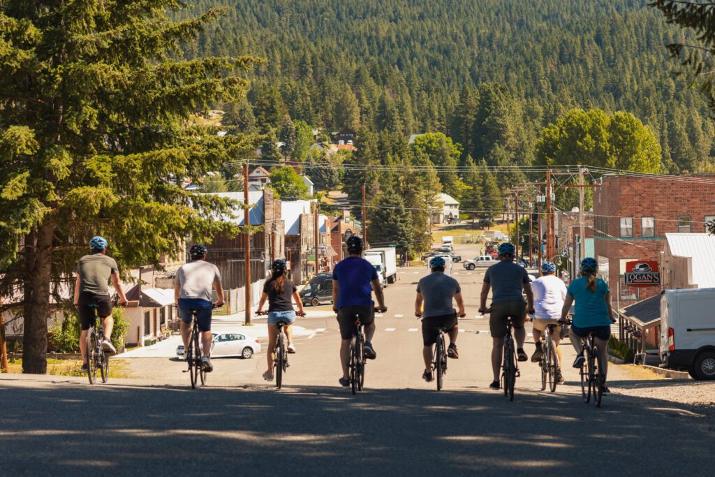 A group of eight people ride bicycles down a street toward a small town surrounded by trees and hills on a sunny day. The riders are wearing helmets and casual clothes.