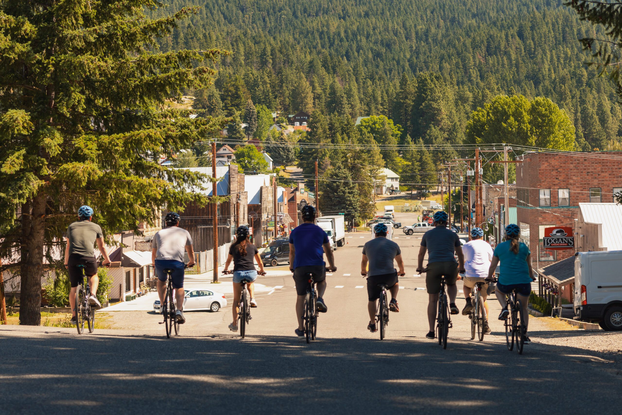 A group of eight people ride bicycles down a street toward a small town surrounded by trees and hills on a sunny day. The riders are wearing helmets and casual clothes.