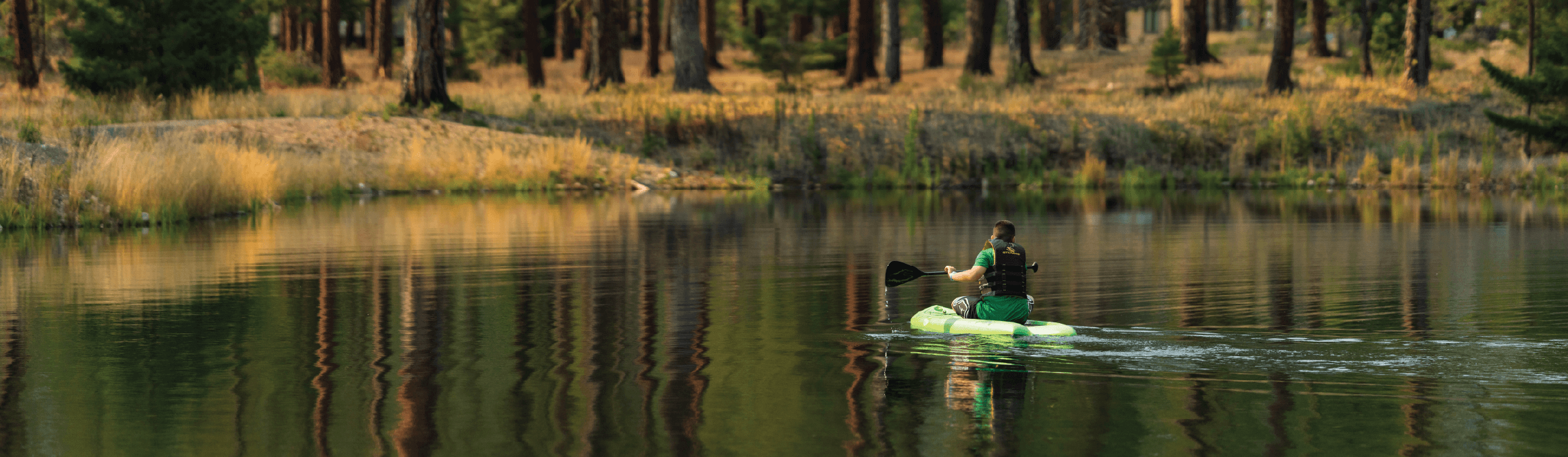 A person paddles a green kayak across a calm lake surrounded by trees and grassy shores, with forest scenery reflected in the water.