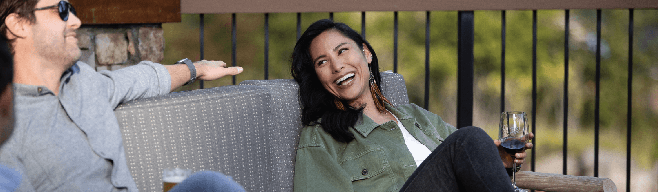 A woman smiles and laughs while sitting on an outdoor chair, holding a glass of wine. A man sitting nearby is talking to her. The setting appears relaxed, with a railing and greenery in the background.