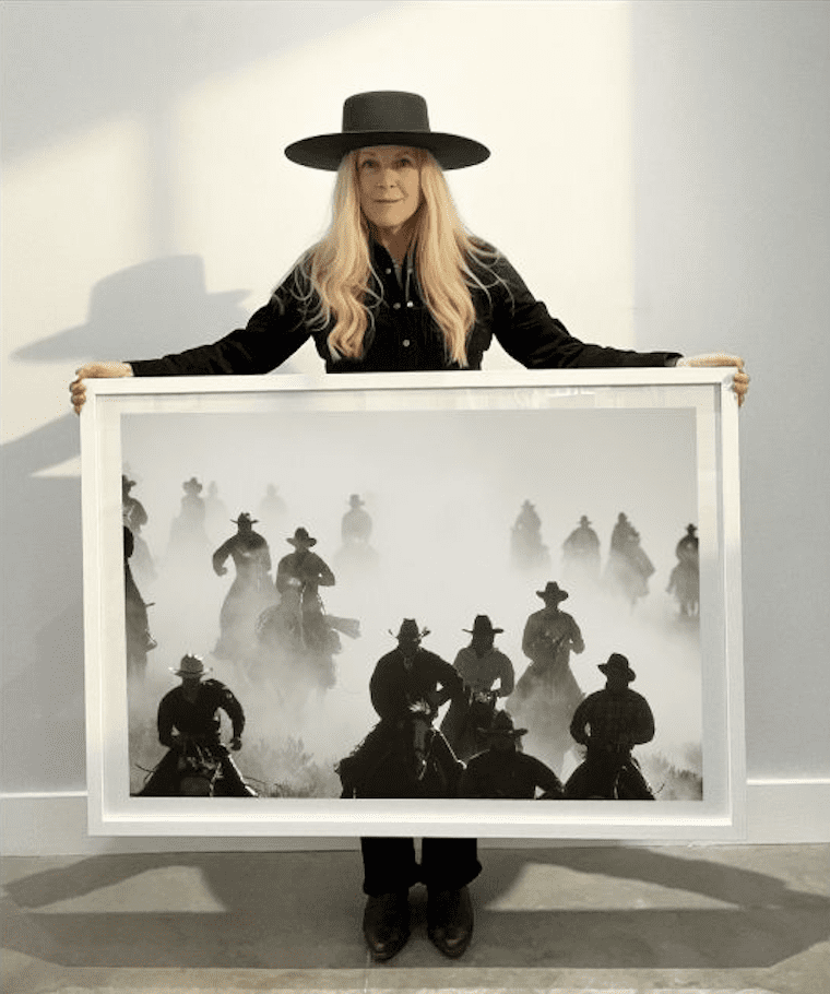 A woman in a black outfit and wide-brimmed hat holds a framed black-and-white photograph of a group of cowboys on horseback, riding through dust.