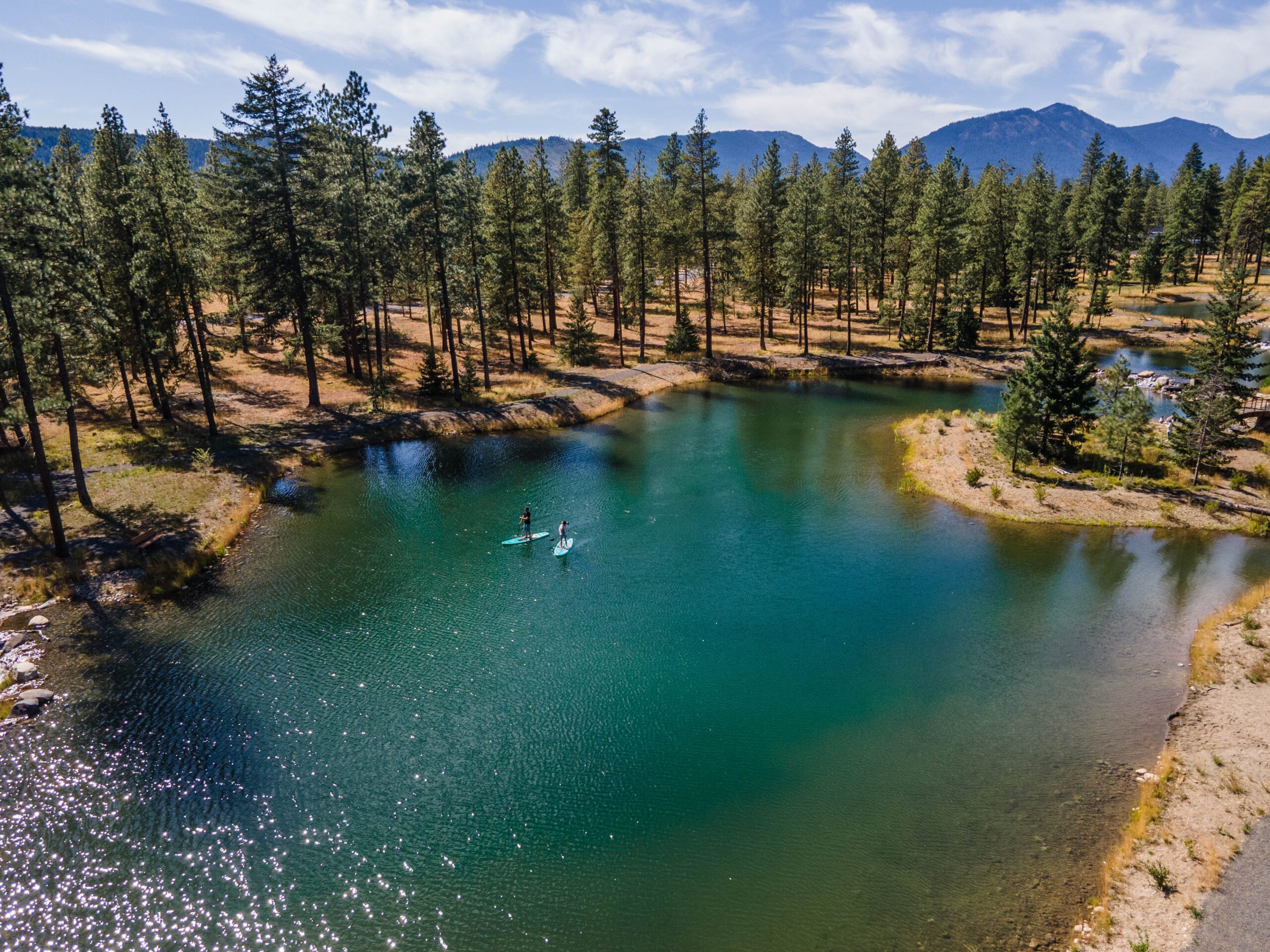 Aerial view of two people paddleboarding on a small, calm lake surrounded by pine trees, with mountains visible in the background under a partly cloudy sky.