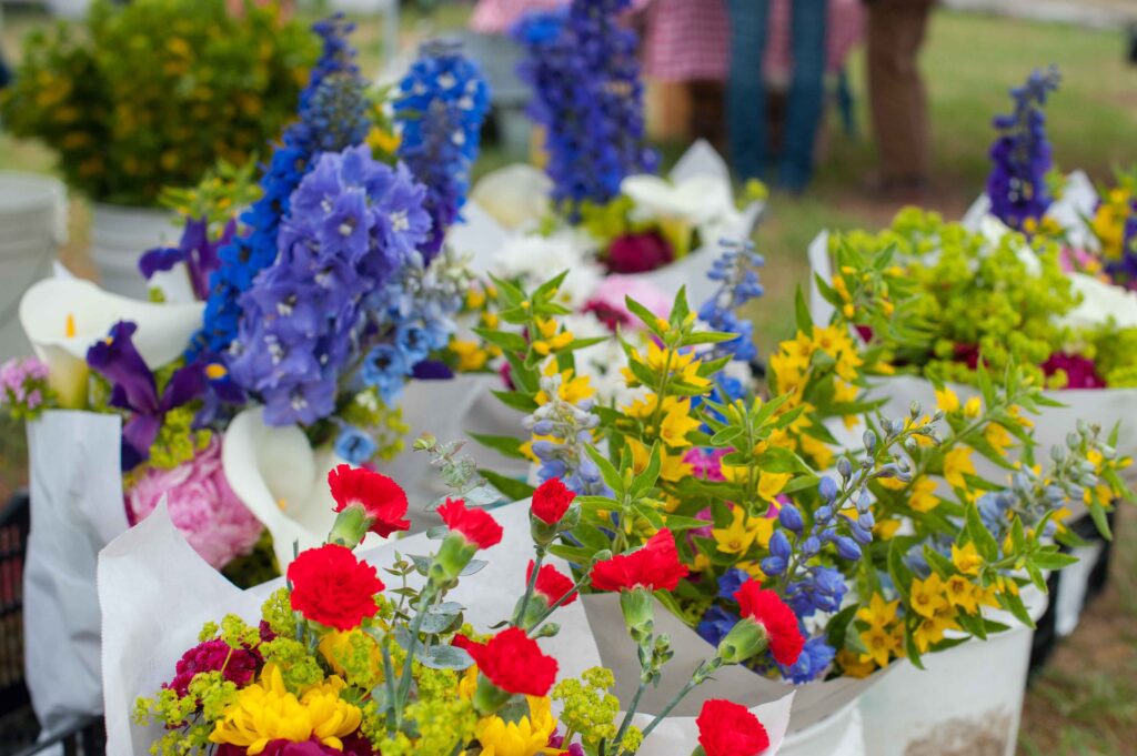 Bouquets of colorful flowers, including red, yellow, blue, and purple blooms, are wrapped in white paper and displayed outdoors, with blurred people and greenery in the background.